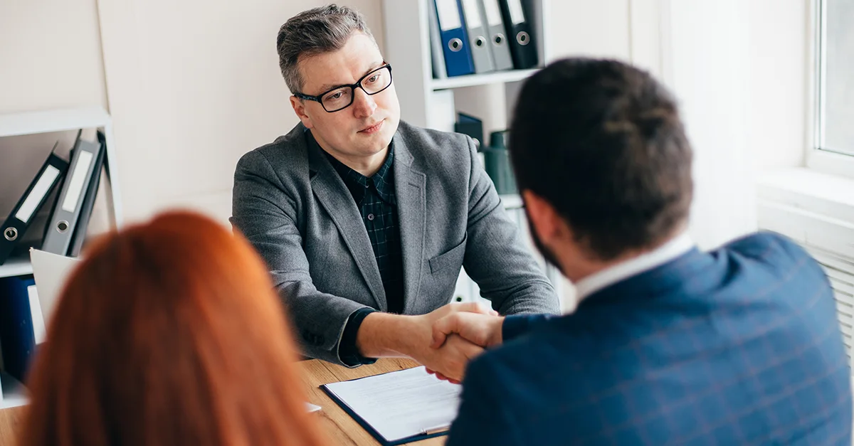 Two coworkers shaking hands across a desk during a meeting, with another person seated nearby and office folders and documents visible in a bright workspace.