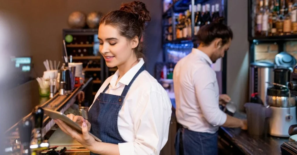 Two cafe workers, one using an ipad while another prepares coffee in the background