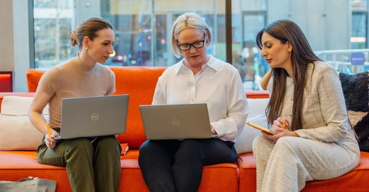 3 female HR colleagues sat on an orange sofa with laptops discussing compliance