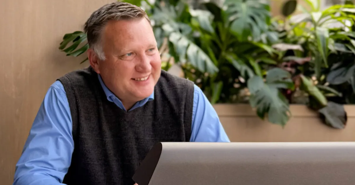 Man with grey vest over blue long sleeve shirt looks out into the distance smiling over his work laptop.