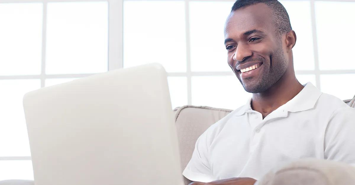 Person seated on a sofa indoors, using a laptop placed on the lap, with soft natural light coming from a window in a minimal home setting.