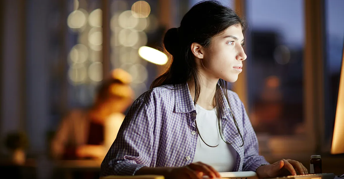 Person seated at a desk indoors at night, working on a laptop near a window, with warm desk lighting and a softly blurred background showing city lights.