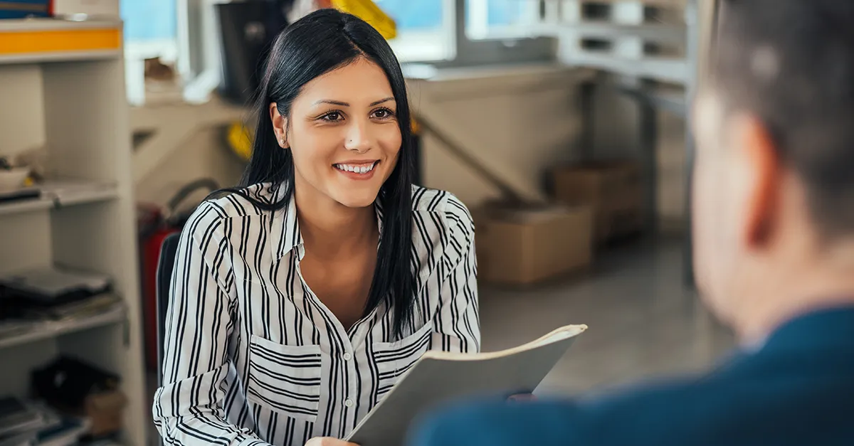 Person seated indoors facing another individual, holding a tablet while engaged in a discussion, with shelves, boxes, and office workspace elements visible in the background.