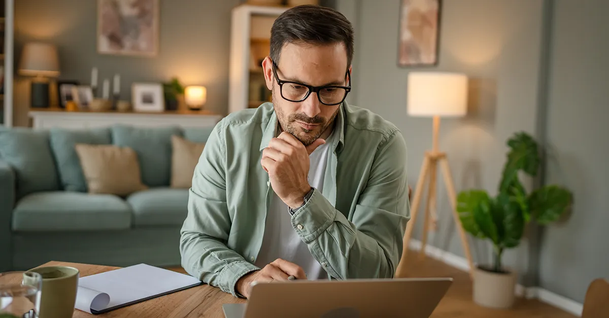 Person wearing eyeglasses seated at a desk in a home office, resting hand near chin while looking at a laptop, with a sofa, lamp, and indoor plant visible in a softly lit background.