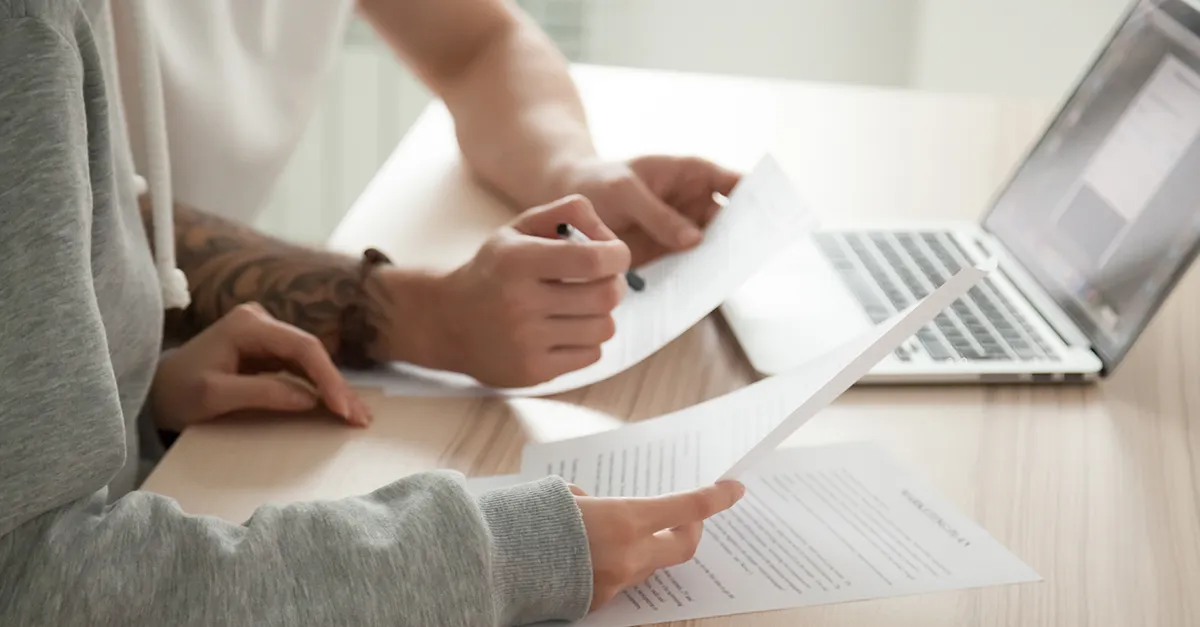 Two people seated at a table, reviewing printed documents together while using a laptop, with hands pointing at pages and a clean, well-lit workspace visible.