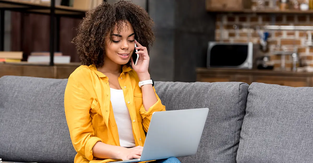 Person seated on a sofa at home, holding a smartphone to the ear while using a laptop on the lap, with a modern interior and soft indoor lighting in the background.