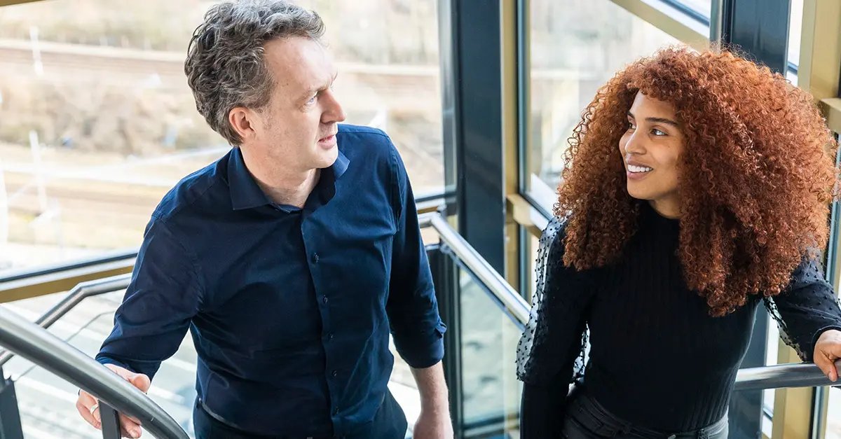 Two coworkers walking side by side indoors in a modern office corridor with large windows, engaged in conversation, with natural light illuminating the space.