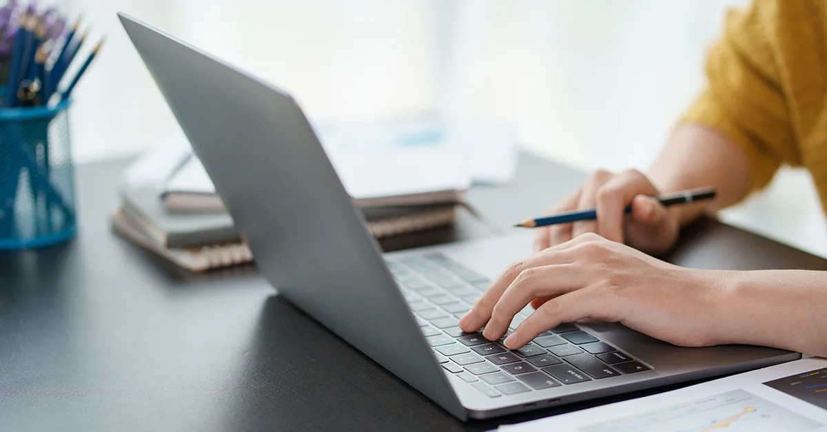 Close-up of hands typing on a laptop keyboard while holding a pen, with a notebook and desk accessories visible in a clean, well-lit workspace.
