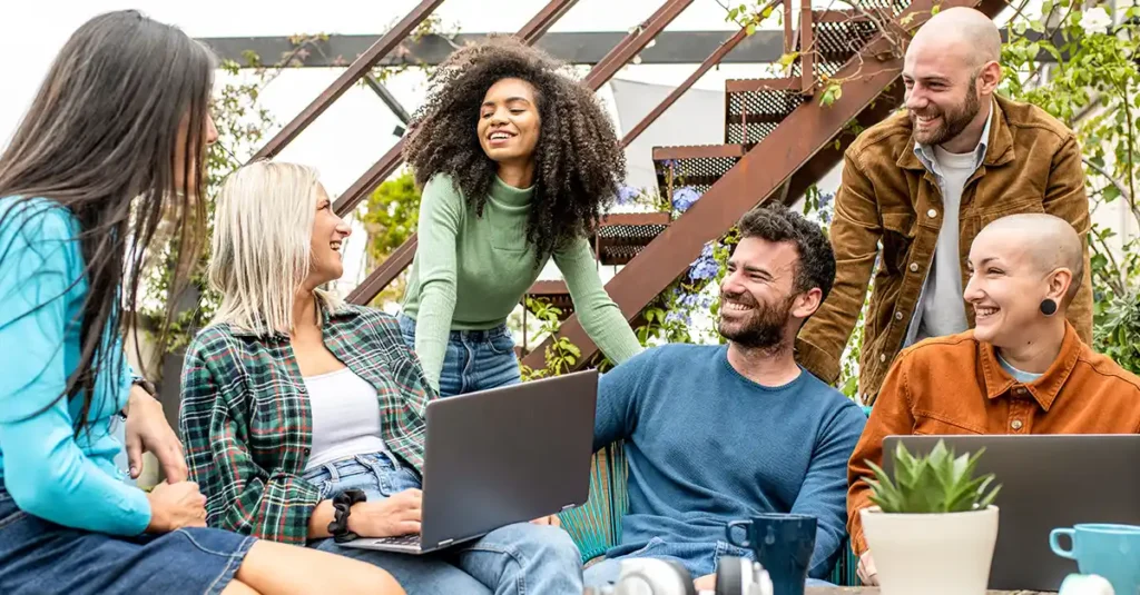 Group of coworkers gathered outdoors near a staircase, collaborating around a laptop while seated and standing together, with plants and an industrial-style background visible.