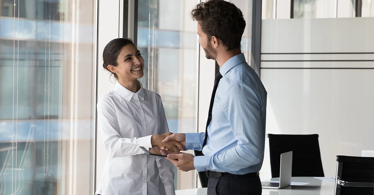 Two coworkers standing indoors in a modern office environment, shaking hands near a glass wall, with a bright workspace and minimal background elements visible.