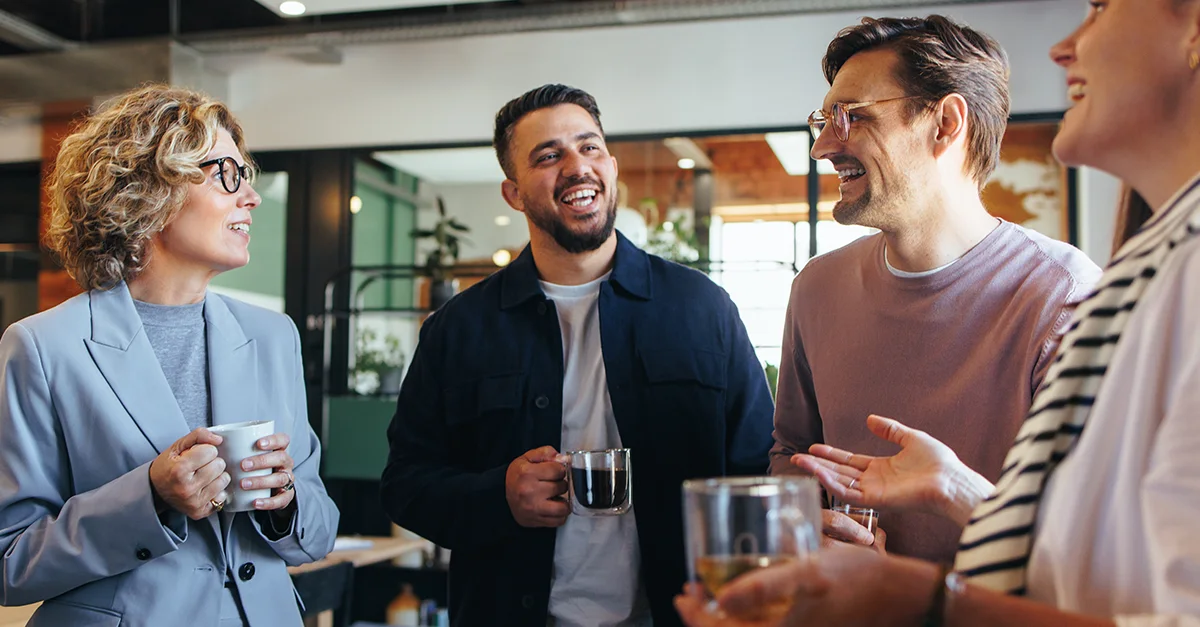 Group of coworkers standing together in a modern office, holding mugs and engaging in a casual conversation, with glass walls and workspace elements visible in the background.