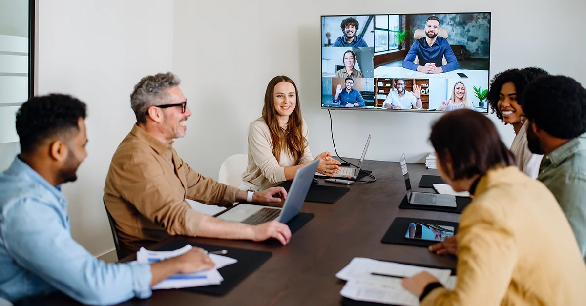 Group of coworkers seated around a table in a modern office, participating in a video conference displayed on a wall-mounted screen, with laptops, documents, and meeting materials on the table.