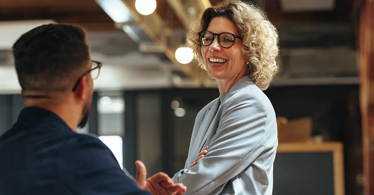 Two professionals shaking hands in a modern office setting, with one holding a pen and wearing glasses, and a blurred workspace background with warm lighting.