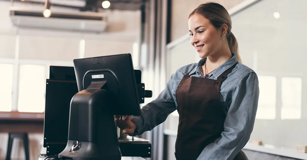 Person standing behind a counter, operating a point-of-sale system on a touchscreen terminal, wearing an apron in a bright indoor retail or café setting.
