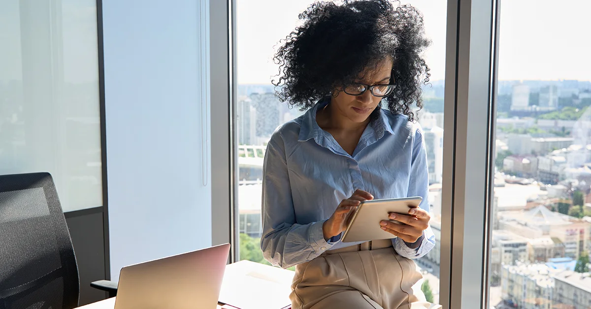 Person standing near a large window holding a tablet and looking at the screen, with a laptop on a nearby table and city buildings visible through the glass in a bright office setting.
