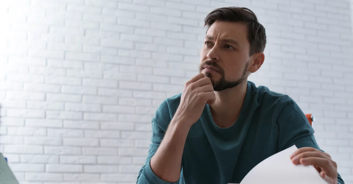 Person seated at a desk holding a sheet of paper and resting a hand on the chin while looking forward, with a white brick wall in the background and soft indoor lighting.