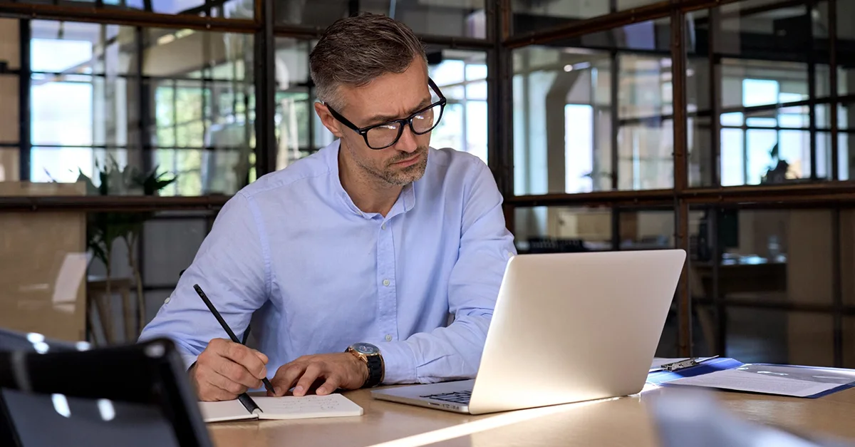 Person seated at a desk writing notes on paper while using a laptop, with documents, a phone, and a bright office interior with glass partitions in the background.