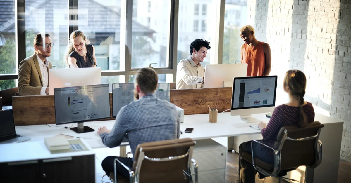 Group of coworkers seated and standing around desks in a bright open-plan office, working on desktop computers and laptops while interacting across workstations, with large windows and glass partitions in the background.