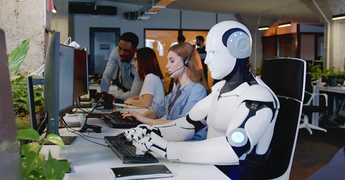 Humanoid robot seated at a desk using a computer alongside a row of people wearing headsets and working on desktop setups in a modern office environment, with monitors, keyboards, and large windows visible in the background.